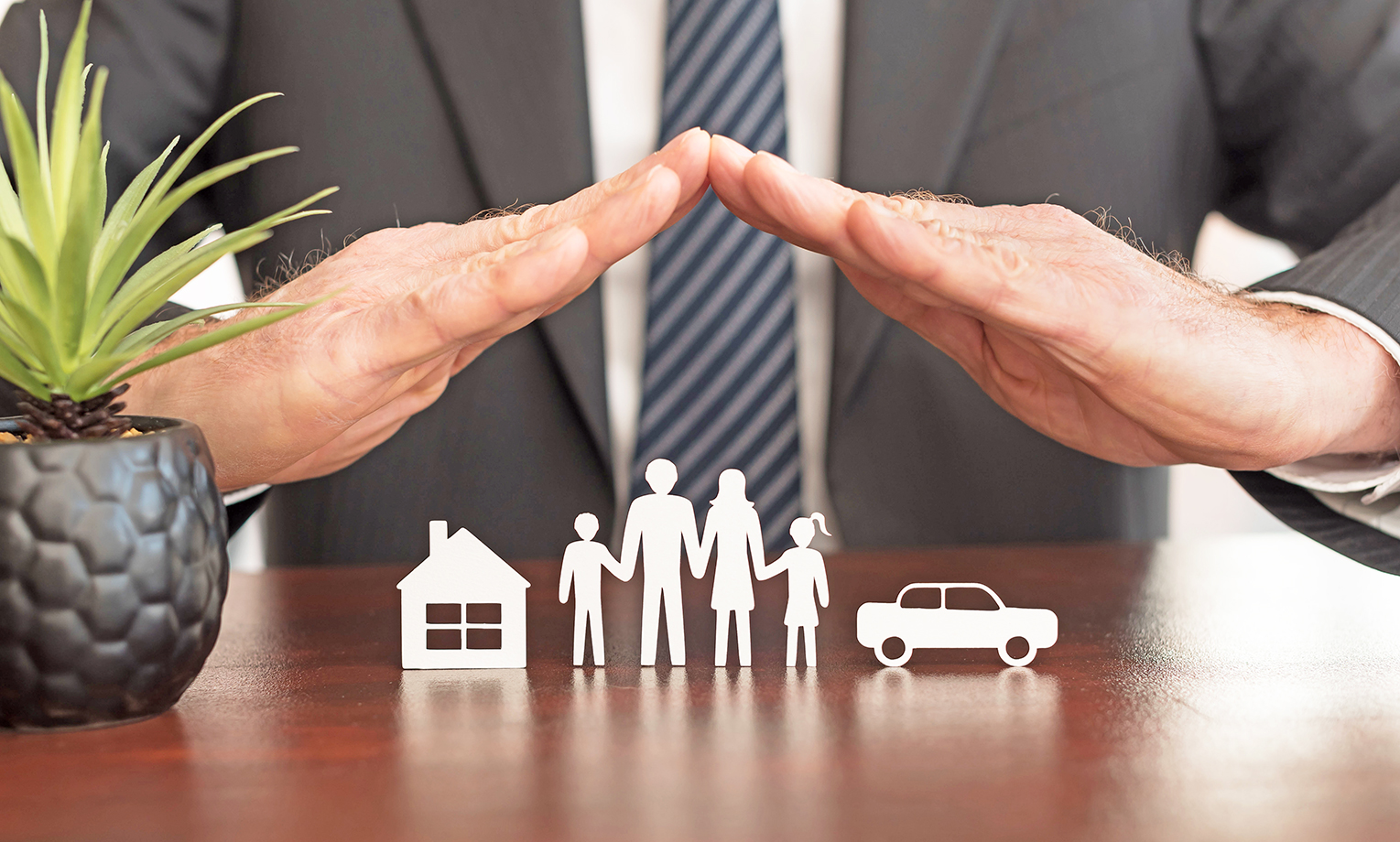 Ein Versicherungsberater schützt symbolisch eine Familie, ein Haus und ein Auto mit seinen Händen, was für finanzielle Sicherheit durch Lebensversicherung und Altersvorsorge steht.