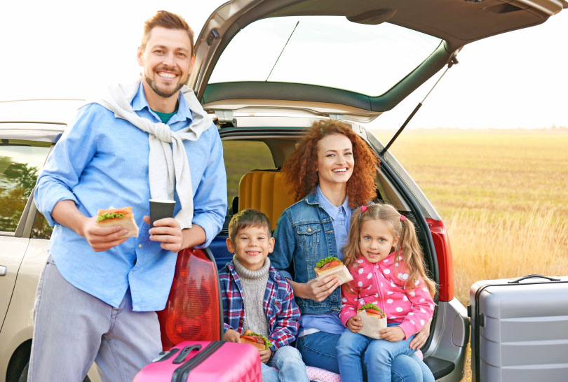Eine fröhliche Familie macht eine Pause auf ihrer Reise, während sie an ihrem offenen Kofferraum sitzt und gemeinsam Sandwiches isst. Im Hintergrund erstreckt sich eine weite Landschaft, und neben dem Auto stehen Koffer, die auf eine längere Reise hindeuten. Dank der richtigen Reiseversicherung genießen sie ihre Fahrt sorgenfrei und mit voller Sicherheit.