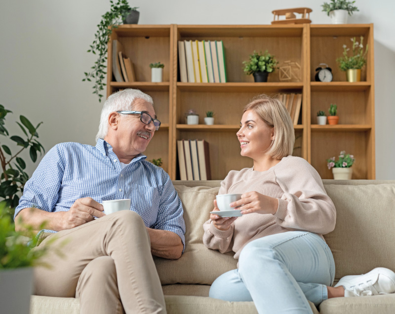 Ein älterer Vater mit grauen Haaren und Brille sitzt entspannt auf dem Sofa und spricht mit seiner Tochter. Sie besprechen in ruhiger Atmosphäre, wie es mit der Wohnung und der Rente weitergeht, um eine gute Lösung für die Zukunft zu finden.