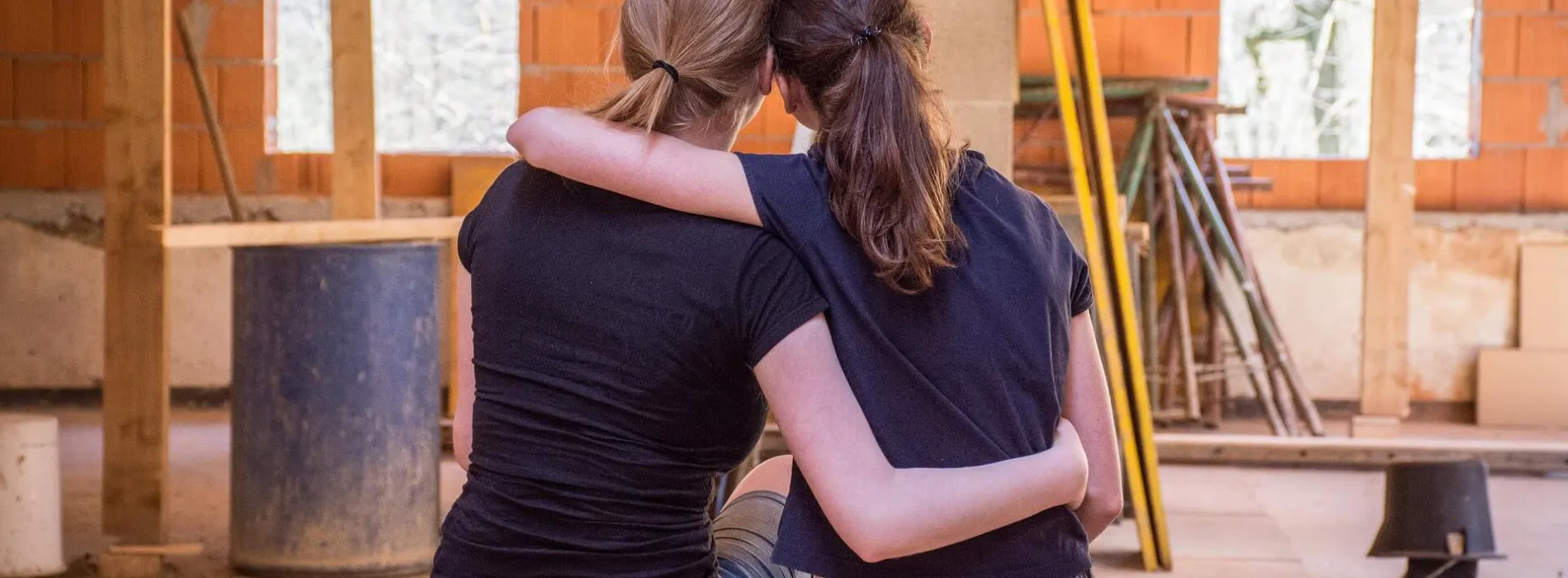 Zwei junge Frauen sitzen Arm in Arm auf einem Holzbalken in einem Rohbau und blicken auf die Baustelle ihres zukünftigen Zuhauses. Im Hintergrund sind unverputzte Ziegelwände, Fensteröffnungen und Baumaterialien zu sehen. Das Bild symbolisiert emotionale Verbundenheit und Verantwortung beim Hausbau. Eine Bauherrenhaftpflichtversicherung in Leipzig schützt vor finanziellen Risiken während der Bauphase.