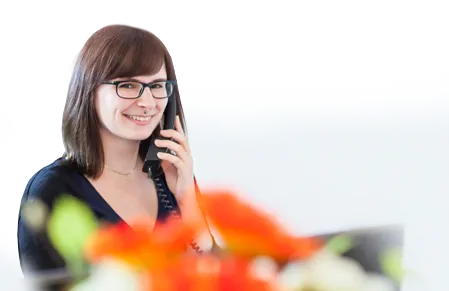 Freundliche junge Frau mit dunklen Haaren und Brille, die am Telefon lächelt. Sie trägt ein dunkles Oberteil und wirkt sympathisch und professionell. Im Vordergrund sind unscharfe Blüten in leuchtenden Farben zu sehen, die eine einladende Atmosphäre schaffen. Das Bild symbolisiert die Möglichkeit, einen Beratungstermin zur Versicherung bei der unabhängigen Finanz- und Versicherungsberatung in Leipzig zu vereinbaren.