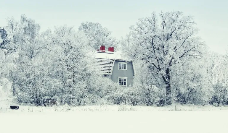 Ein Einfamilienhaus ist von verschneiten Bäumen und tiefem Winterweiß umgeben. Das Dach ist mit Schnee bedeckt, die Landschaft wirkt idyllisch, aber auch potenziell gefährlich durch Frost- und Schneelasten. Das Bild unterstreicht die Bedeutung einer zuverlässigen Immobilienversicherung in Leipzig, um Wohngebäude ganzjährig gegen Witterungsschäden abzusichern.