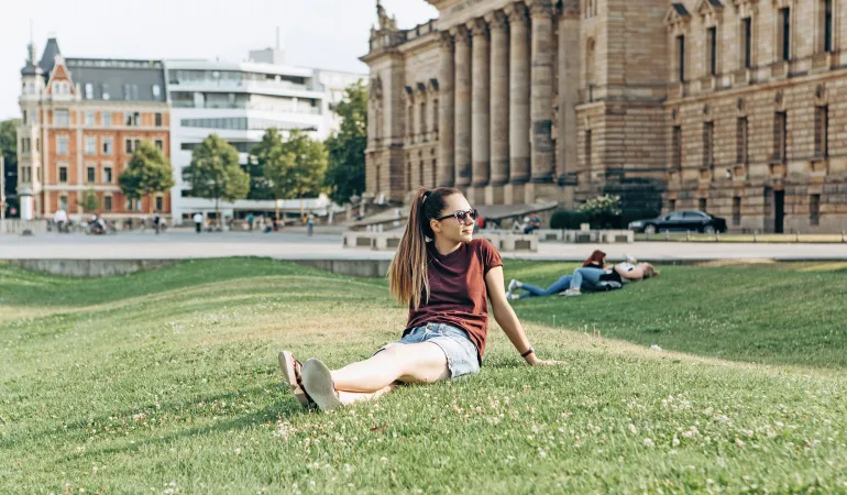 Eine Person sitzt entspannt auf einer Wiese vor einem historischen Gebäude in Leipzig. Diese Szene symbolisiert das Studentenleben, inklusive der Wichtigkeit einer Krankenversicherung für Studenten in Leipzig.