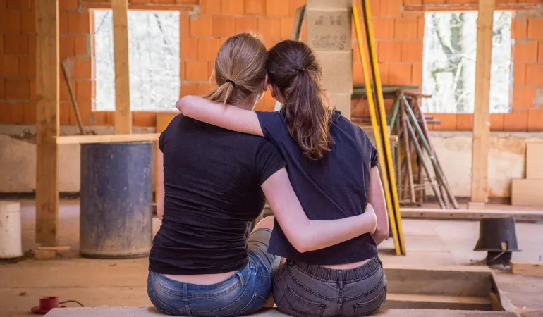 Zwei junge Frauen sitzen Arm in Arm auf einem Holzbalken in einem Rohbau und blicken auf die Baustelle ihres zukünftigen Zuhauses. Im Hintergrund sind unverputzte Ziegelwände, Fensteröffnungen und Baumaterialien zu sehen. Das Bild symbolisiert emotionale Verbundenheit und Verantwortung beim Hausbau. Eine Bauherrenhaftpflichtversicherung in Leipzig schützt vor finanziellen Risiken während der Bauphase.
