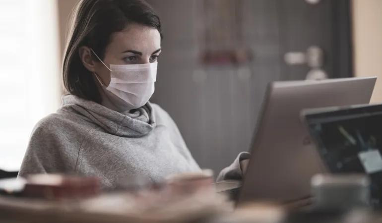 Frau mit medizinischer Maske arbeitet im Homeoffice am Laptop – Erwerbsunfähigkeitsversicherung Leipzig schützt bei gesundheitlicher Arbeitsunfähigkeit