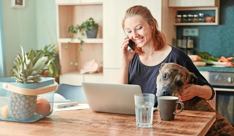 Frau informiert sich über Haftpflichtversicherung am Laptop, telefoniert und streichelt ihren Hund in gemütlicher Küche