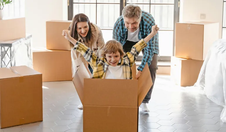 Familie beim Umzug: lachendes Kind sitzt in einem Karton, Eltern schieben weitere Umzugskartons im neuen Zuhause — Symbolbild für Hausratversicherung.