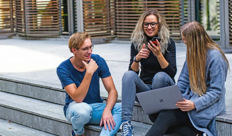 Drei junge Menschen sitzen auf einer Treppe, nutzen Smartphone und Laptop, und sprechen über ihre finanzielle Zukunft mit Fokus auf Altersvorsorge, Rente und mögliche Sonderzulagen