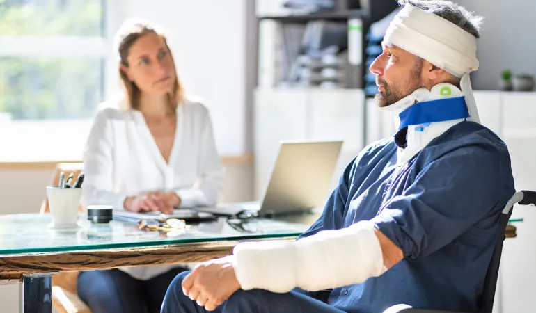 Verletzter Mann mit Gips und Halskrause im Rollstuhl bei einem Beratungsgespräch über Absicherung im Krankheitsfall und Unfallfolgen