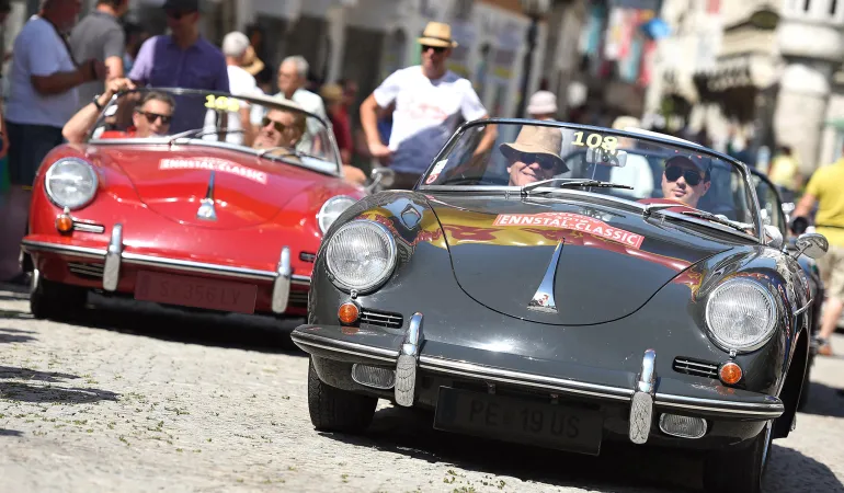 Zwei Porsche-Oldtimer fahren bei einer klassischen Oldtimer-Rallye durch eine belebte Altstadt.