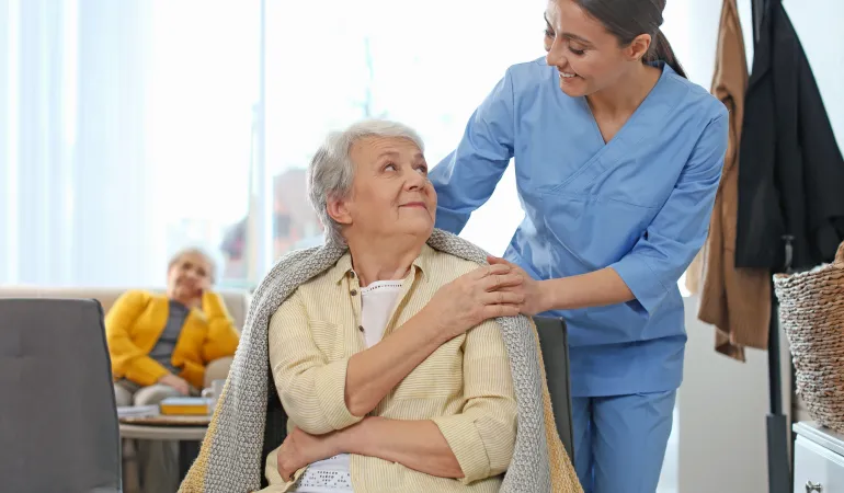 Eine freundliche Pflegerin in blauer Arbeitskleidung kümmert sich liebevoll um eine ältere Dame, indem sie ihr fürsorglich eine Decke umlegt. Die Seniorin sitzt entspannt und blickt dankbar nach oben. Im Hintergrund ist eine weitere ältere Frau in einem gemütlichen Wohnbereich zu sehen. Die Szene symbolisiert professionelle Pflege und Betreuung im Alter. Pflegekosten in Leipzig sind ein wichtiges Thema für Senioren und ihre Angehörigen, um die bestmögliche Versorgung sicherzustellen.