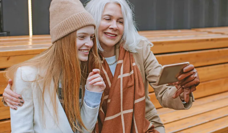 Ältere Frau und junge Frau sitzen gemeinsam auf einer Bank, lächeln beim Blick aufs Smartphone – Symbol für Generationen und gemeinsame Altersvorsorge bei Rente ab 73.