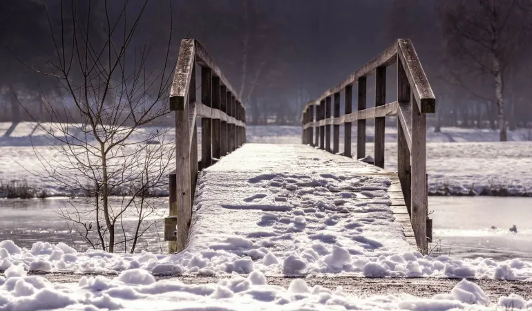 Verschneite Holzbrücke über zugefrorenem Gewässer in winterlicher Landschaft – Symbol für Sicherheit auf dem Eis und passenden Versicherungsschutz
