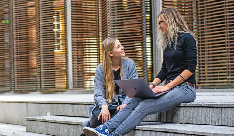 Zwei junge Erwachsene sitzen auf einer Treppe und unterhalten sich über Versicherungsrabatte – eine nutzt ein Laptop