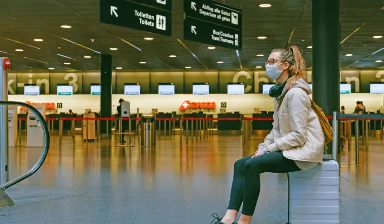 Reisende mit Mund-Nasen-Maske sitzt auf ihrem Koffer im Flughafen-Check-in-Bereich und wartet auf den Flug in den Urlaub ins Hotel unter Virus-Schutzauflagen